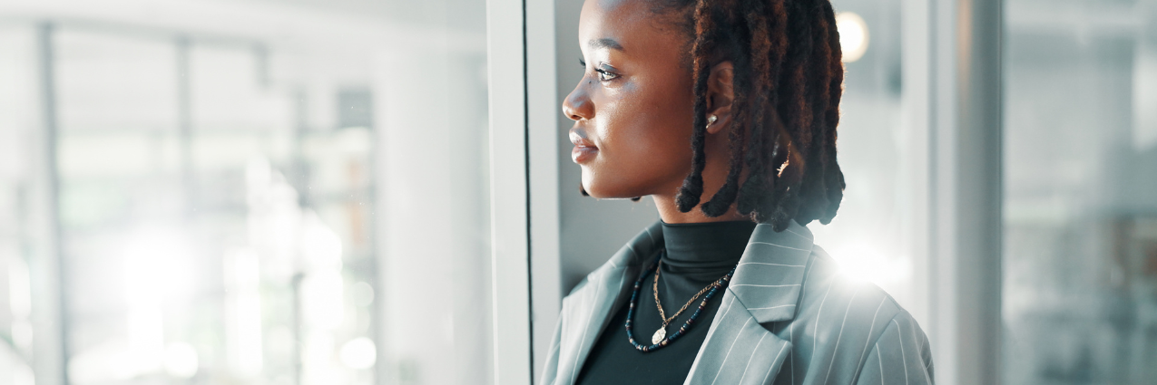 Black businesswoman looking out of an office window, representing leadership, reflection, and women's empowerment in the workplace